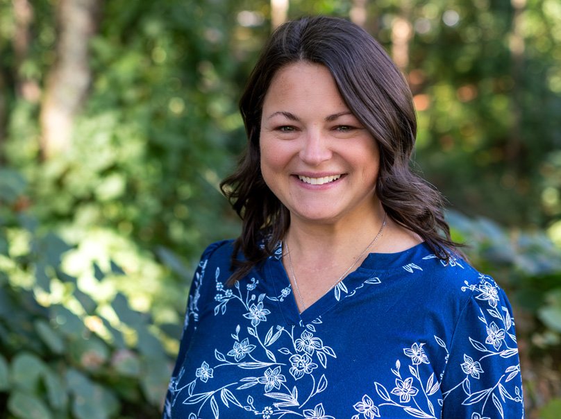 Katie Taylor smiling with shoulder-length dark hair wearing a blue floral blouse, standing outdoors with greenery in the ba
