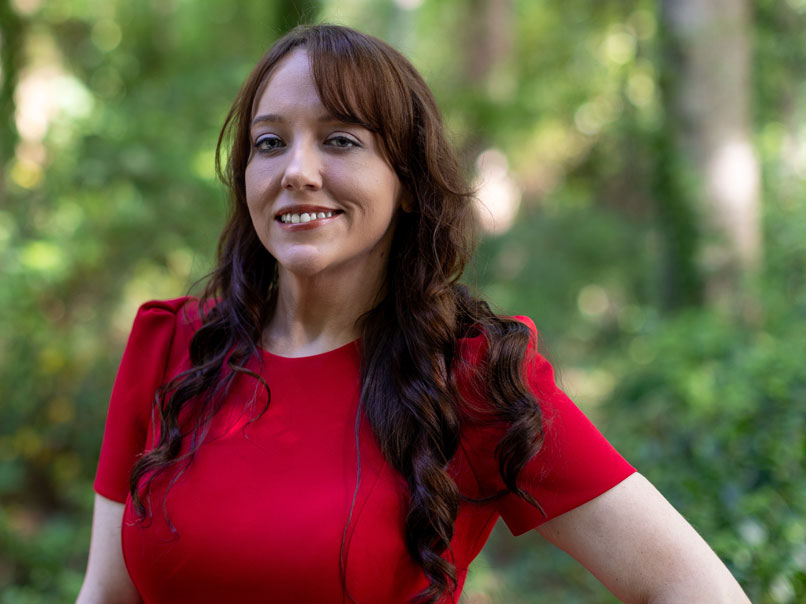 Woman in a red top standing outdoors with a blurred wooded background. Click to view bio.
